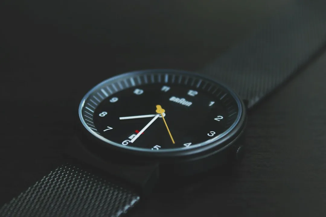 A close-up view of a black analog wristwatch with a textured mesh strap resting on a dark table.