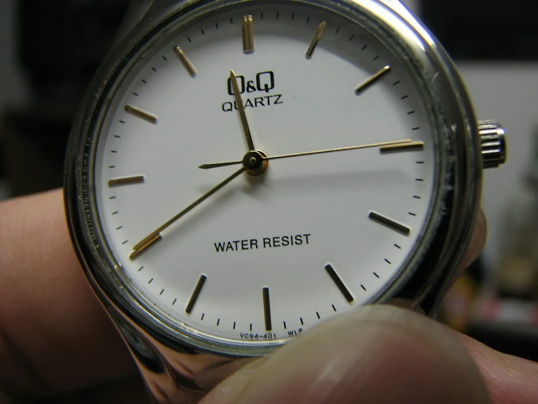A close-up of a silver wristwatch with 'WATER RESIST' printed on the dial being held by fingers.