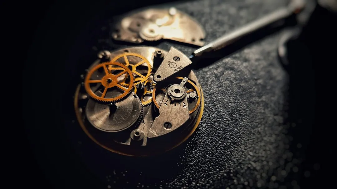 A disassembled mechanical watch movement with gears and components laid out on a dark textured surface.