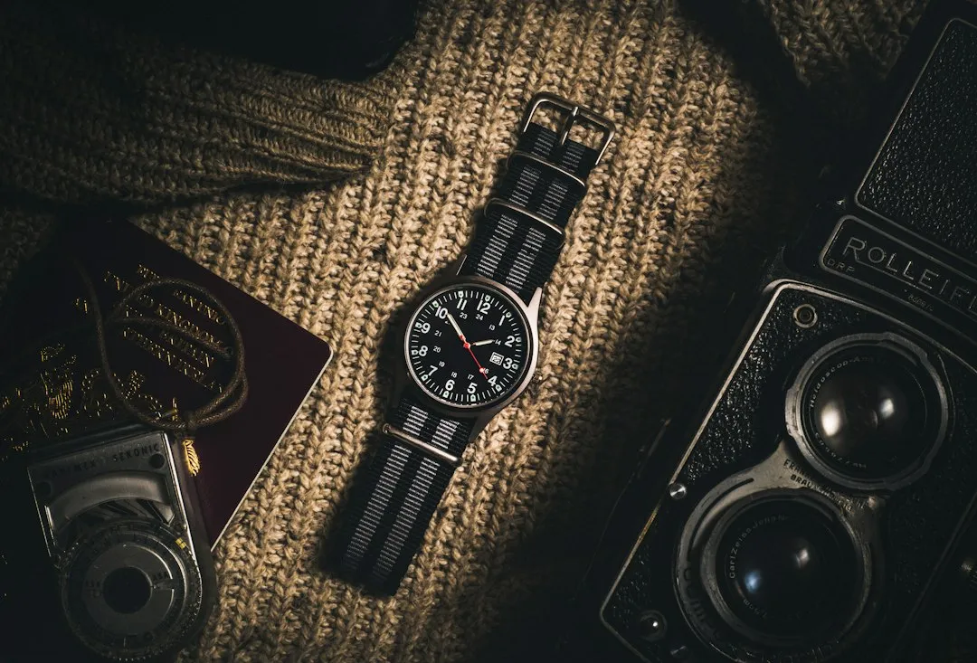 A wristwatch with a striped NATO strap lying on a knitted sweater next to a passport and camera.