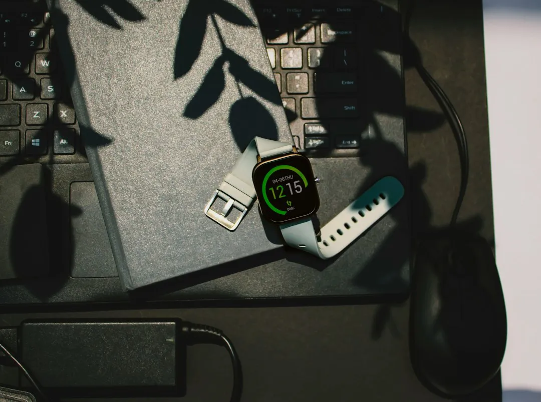 A smartwatch showing 12:15 lies on a grey notebook placed over a laptop keyboard with leaf shadows.