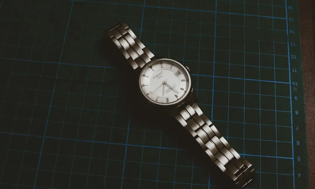 A silver wristwatch with a metal link bracelet lies on a green grid cutting mat.