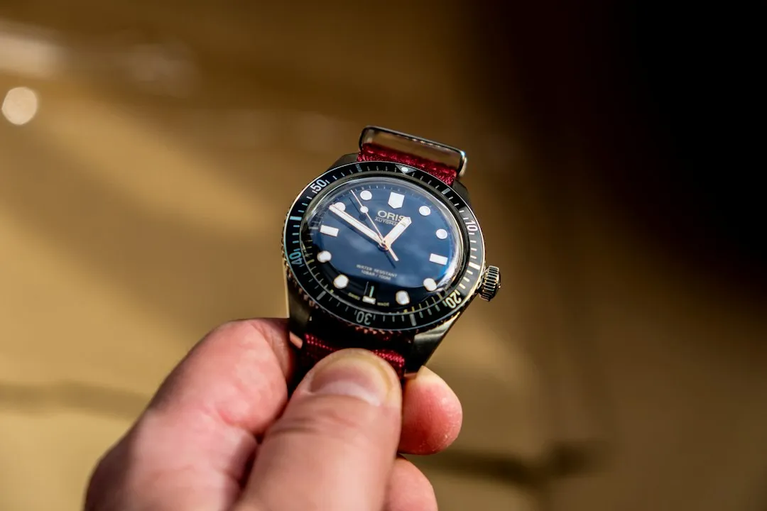 Un primer plano de una mano sosteniendo un reloj Oris de buceo con esfera azul y correa de cuero roja.