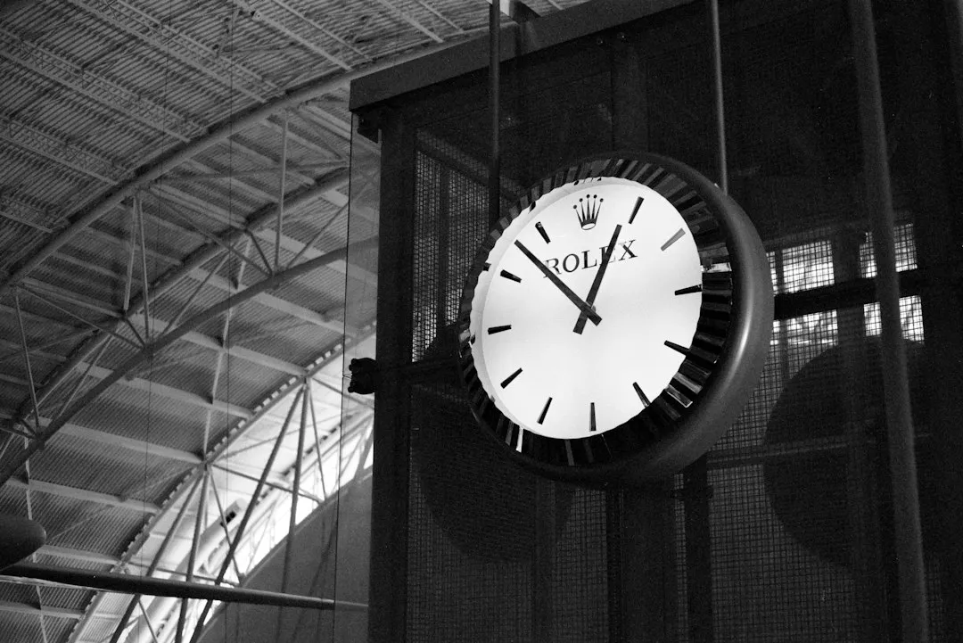 A black and white photo of a large Rolex wall clock hanging from a metal framework indoors.