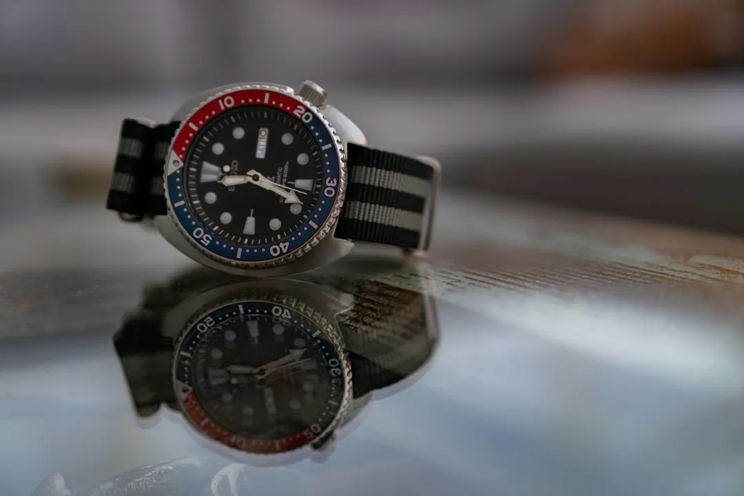 A vintage-style cushion case dive watch with a red and blue bezel rests on a glass surface.