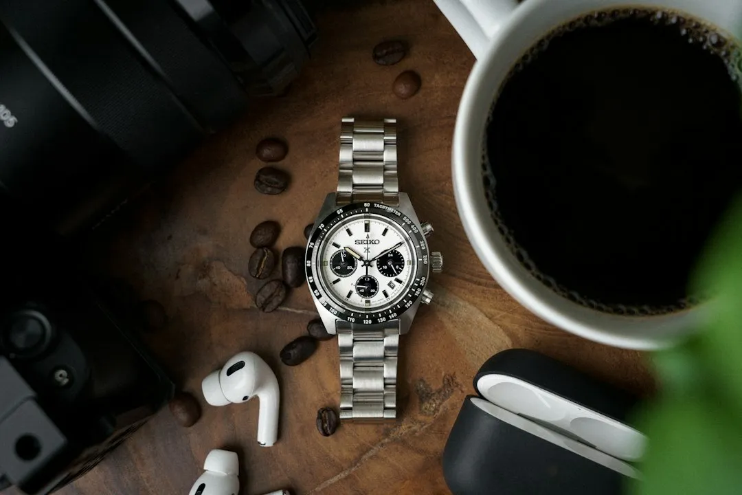 A stainless steel chronograph wristwatch with a white panda dial and black sub-dials rests on a wooden table beside coffee.