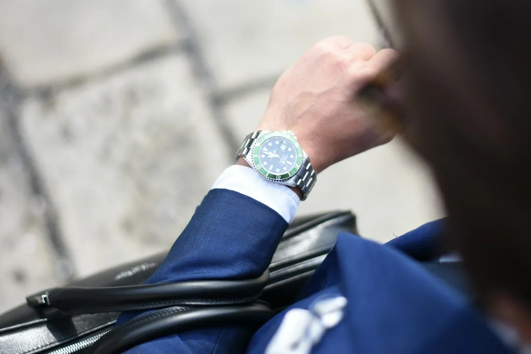 A person wearing a silver wristwatch with a green bezel under a blue suit sleeve cuff.