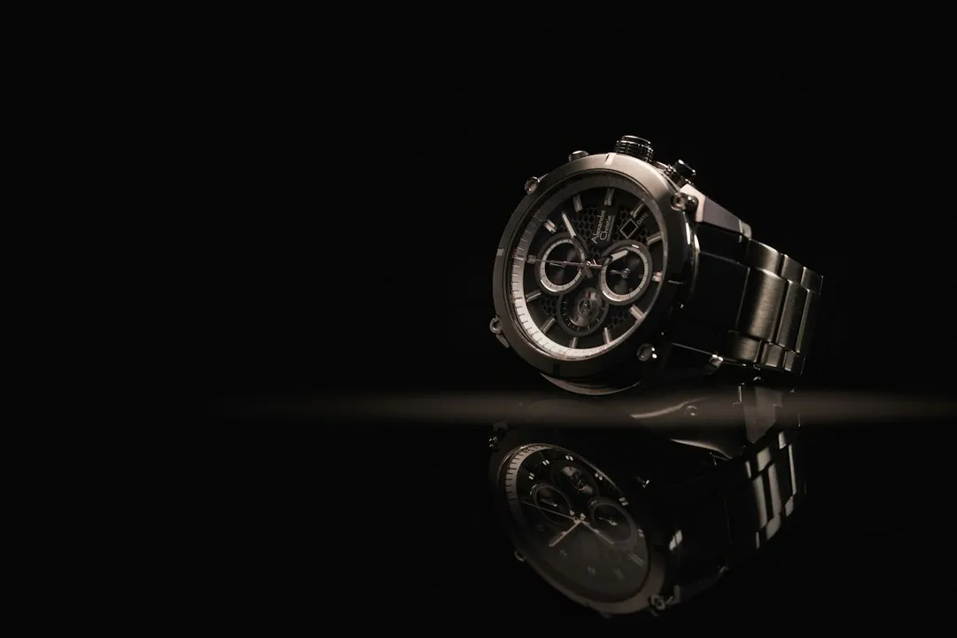 A silver chronograph wristwatch with a metal bracelet sits on a reflective black surface.