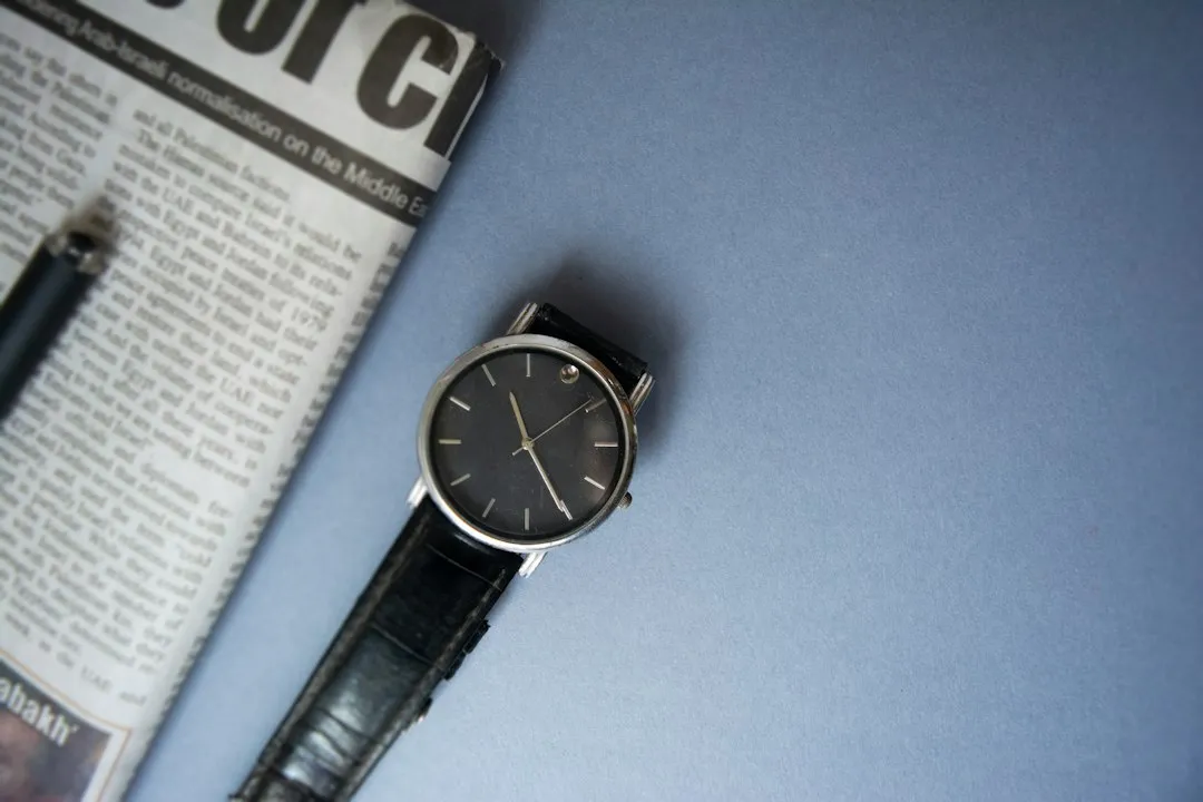 A minimalist wristwatch with a black leather strap rests on a blue surface next to a newspaper.
