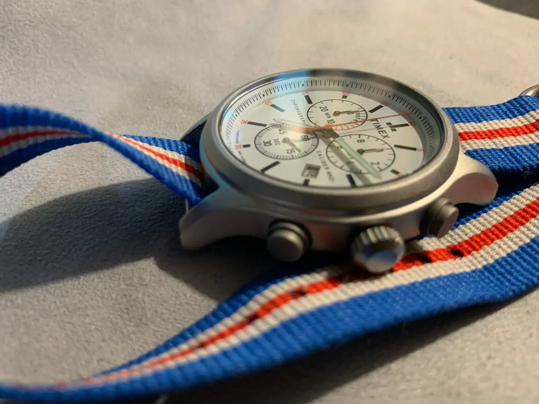 A wristwatch featuring a classic blue, red, and white striped NATO strap resting on a fabric surface.