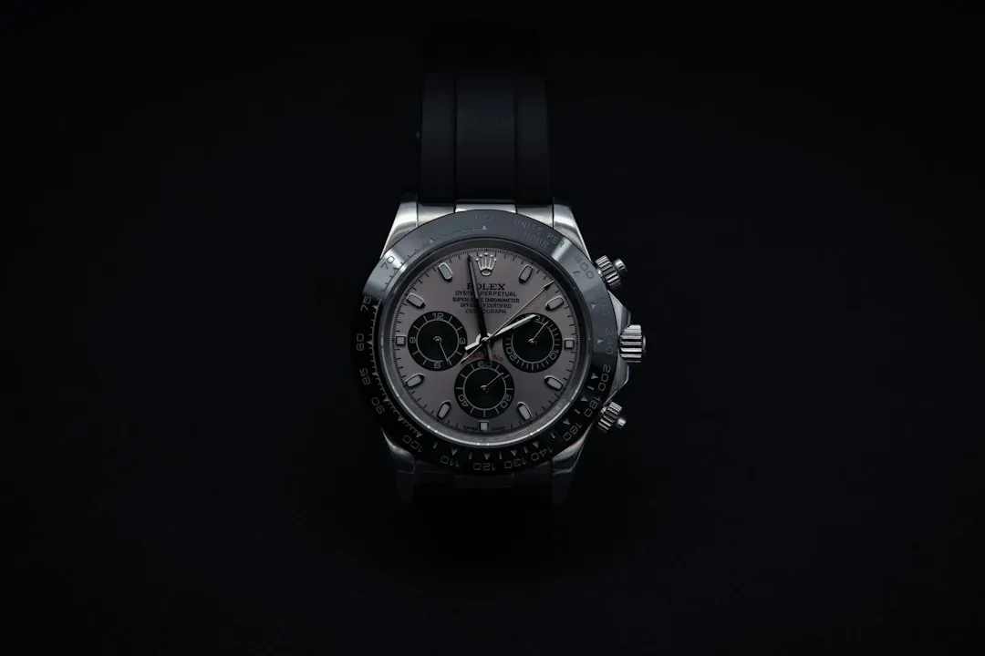 A black and white close-up of a silver Rolex chronograph wristwatch with a black strap on a dark background.