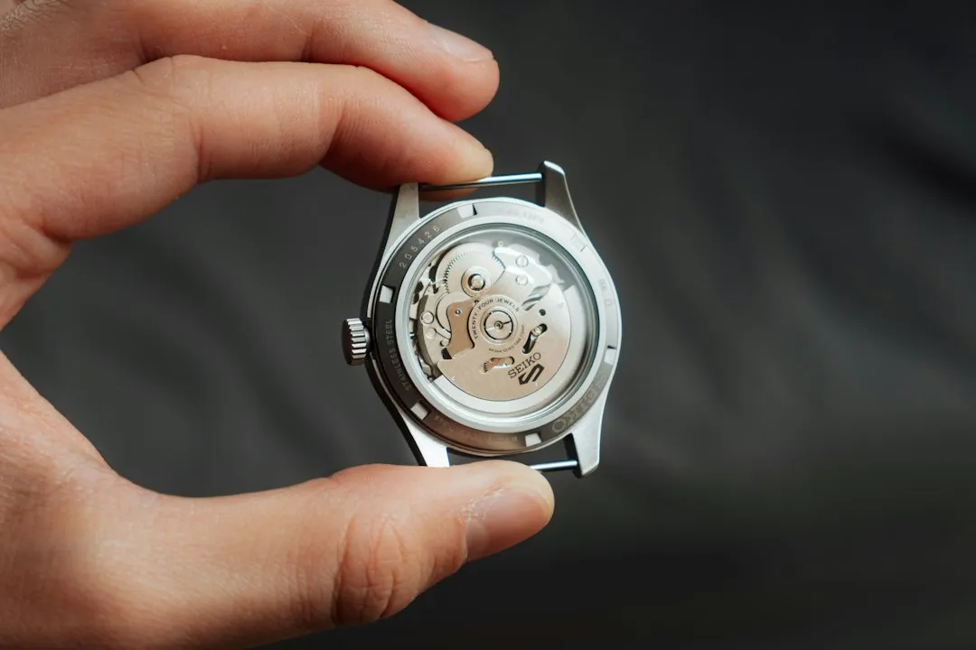 Hands holding a silver watch case back, exposing the gold-tone gears and balance wheel of the movement.