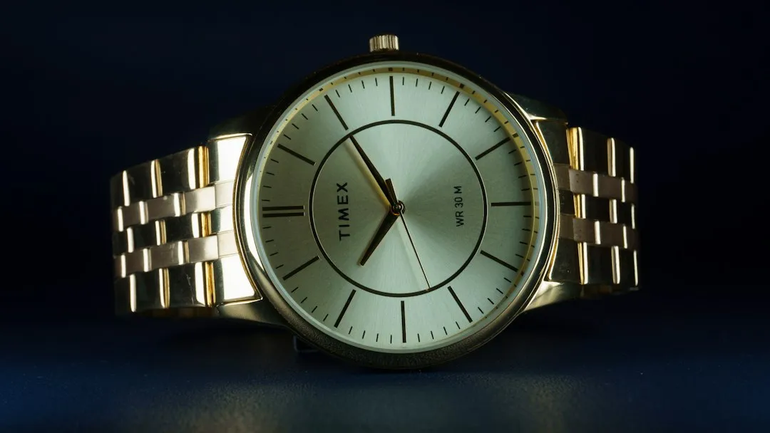 A gold Timex wristwatch with a white dial and linked metal bracelet against a dark background.