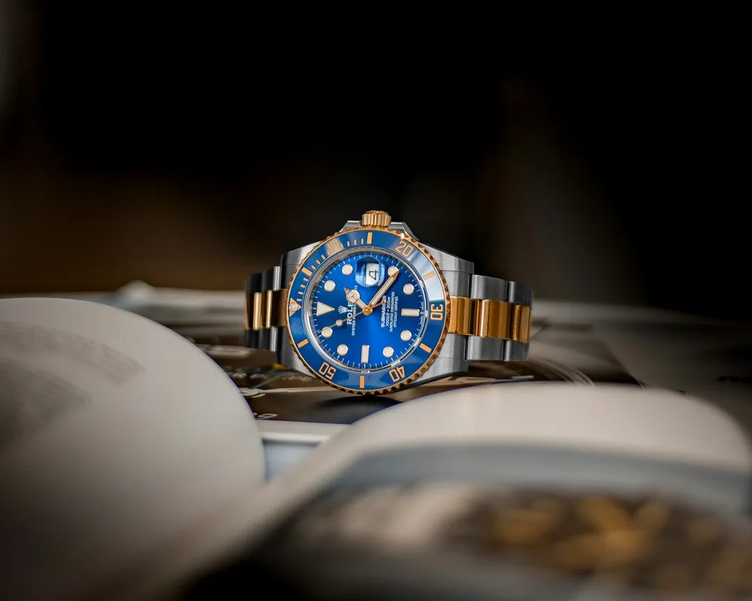 A close-up of a blue dial luxury wristwatch with a two-tone metal bracelet resting on magazines.